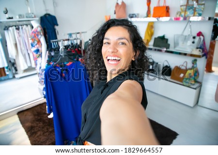 Cheerful excited Latin woman taking selfie near rack with dresses in fashion shop, looking at camera, smiling and laughing. Boutique customer or shop assistant concept