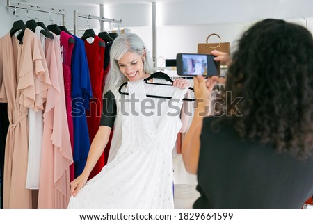 Happy female friends enjoying shopping in clothes store together, holding dress, posing and taking pictures on mobile phone. Consumerism or shopping concept