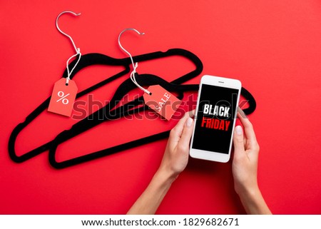 Black Friday Sale concept. Smartphone in female hands with sign "Black Friday" on screen and clothes hangers with tags on red background.