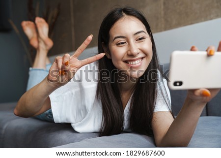 Image of a cheery young woman at home indoors lying on a sofa and taking a selfie by mobile phone with peace gesture