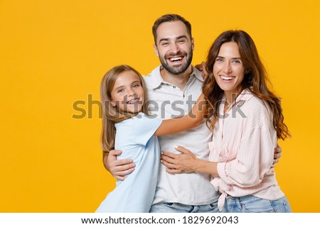 Laughing young happy parents mom dad with child kid daughter teen girl in basic t-shirts hugging looking camera isolated on yellow background studio portrait. Family day parenthood childhood concept