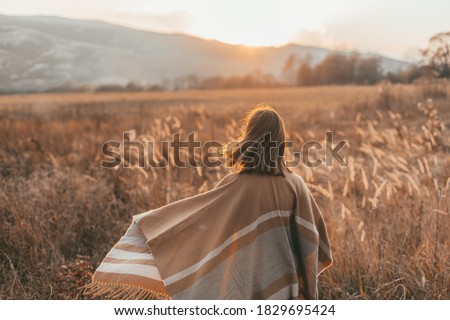 Girl in poncho travel alone in field with a view in sunlight. Warm autumn weather, calm scene. Wanderlust photo series.