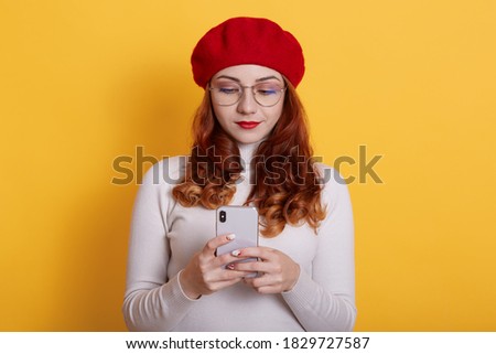 Young brunette Caucasian woman holds modern mobile phone, sends messages, reads funny notification, wears red beret, white shirt and spectacles, poses indoor against yellow wall.