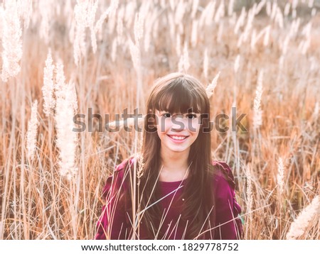 Having fun on Autumn. Warm Fall season nature photo of a cute smiling wide teen girl wearing magenta color sweatshirt at wild fluffy spikes orange and yellow field