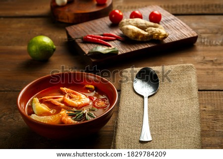 Tom yam soup with shrimp and lime in a brown dish on a wooden table next to a spoon on a napkin and ingredients.