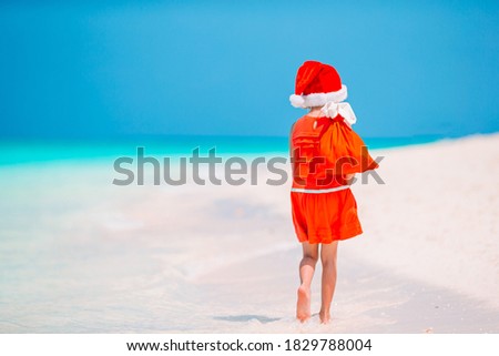Adorable little girl in Santa hat with a bag of gifts on tropical beach