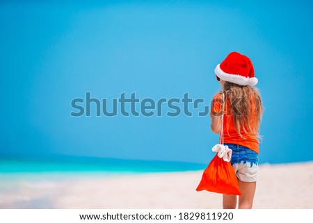 Adorable little girl in Santa hat with a bag of gifts on tropical beach