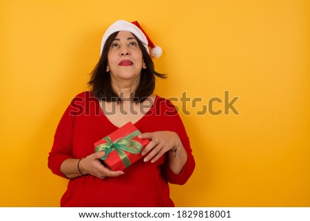 portrait of mysterious charming middle aged woman wearing Christmas hat and red sweater, looking up with enigmatic smile. Beautiful smiling girl looking up standing against orange wall.