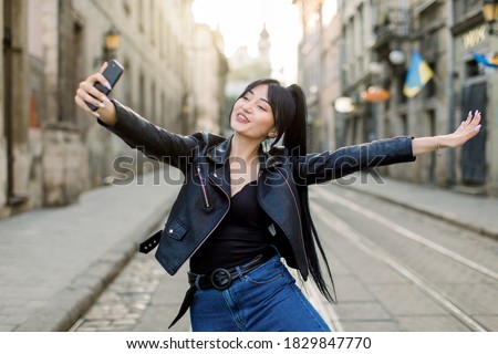 Happy female mixed raced tourist taking selfie on city street, smiling and having fun, enjoying her walk. Young Asian lady in fashionable clothes making selfies photo on the phone