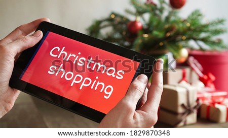 Young woman holding in hands a tablet with text christmas shopping the background of Christmas decor and gifts, close-up. Christmas and New Year shopping on the Internet, payment by credit card.