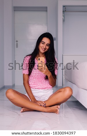 European fit brunette fashion blogger woman in bedroom at home, wearing white denim shorts and cute pink marshmallow shirt, and take photo selfie on mobile phone in mirror.  