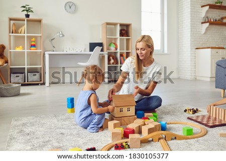 Mom and child at home. Young mother playing with little son teaching him to build toy house. Happy nanny engaging toddler boy in fun activities with wood blocks on warm floor in cozy studio apartment Royalty-Free Stock Photo #1830105377