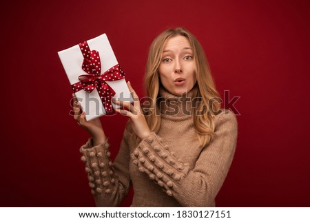 Image of charming young blonde woman in warm sweater smiling and holding gift with red ribbon. Studio shot red background. New Year Women's Day birthday holiday concept