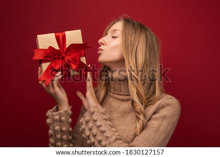 Image of charming young blonde woman in warm sweater smiling and holding gift with red ribbon. Studio shot red background. New Year Women's Day birthday holiday concept