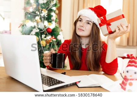 Young woman in red santa hat thinking on something, holding cup with drink and present box, wearing red jumper, sitting next to table and laptop, home office, christmas work time, tree on background