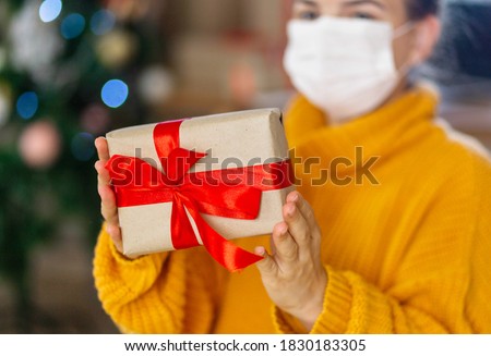 Christmas mask congratulations. Portrait woman wearing yellow sweater in medical mask, giving gift present box with red ribbon,christmas tree bokeh on background Royalty-Free Stock Photo #1830183305