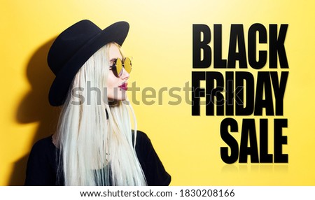 Side portrait of young blonde girl wearing black hat and sunglasses, on background of yellow color with Black Friday Sale text. Holidays concept. 