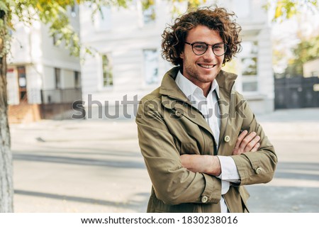 A young successful businessman in eyewear smiling broadly posing outdoors. Male entrepreneur resting in the city street. Smart guy in casual wears spectacles with curly hair walking outside after work Royalty-Free Stock Photo #1830238016