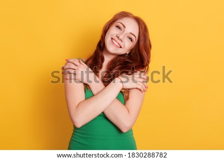 Red haired young beautiful woman looks smiling directly at camera, crossed arms, keeping hands on shoulders, happy girl in green t shirt against yellow wall.