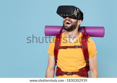 Cheerful traveler young man in yellow casual t-shirt with backpack isolated on blue background studio. Tourist traveling on weekend getaway. Tourism discovering hiking concept. Watching in headset