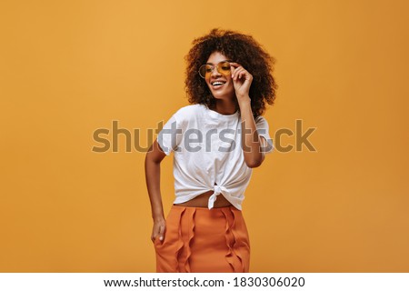 Charming girl with fluffy hair in bright sunglasses poses on isolated backdrop. Lady in white t-shirt and orange skirt smiles on yellow background..