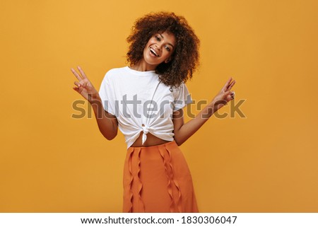 Good-humored woman with wavy hair showing peace sign on isolated background. Modern girl in orange skirt and t-shirt smiling on yellow backdrop..