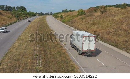 Aerial shot of truck with cargo trailer driving on road in rural environment. Flying over lorry moving along countryside highway. Concept of logistic and delivery goods. Freeway traffic. Back view.