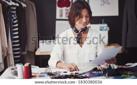 Satisfied fashion designer holding paper looking at sketches sitting at table in clothing atelier