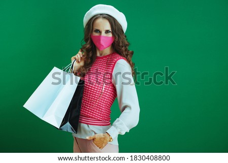 Life during coronavirus pandemic. Portrait of modern woman shopper in pink sleeveless shirt with pink medical mask and shopping bags isolated on green background.