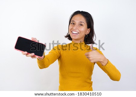 Portrait Young woman with short hair wearing casual yellow sweater isolated over white background, holding in hands cell showing giving black screen thumb up 