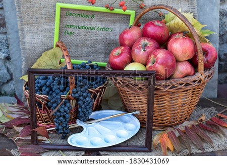 A basket of red apples and a basket of grapes stands on a jute fabric. Nearby lie autumn leaves, an easel, brushes and picture frames. Above the inscription: "December 8 International Artist's Day"
