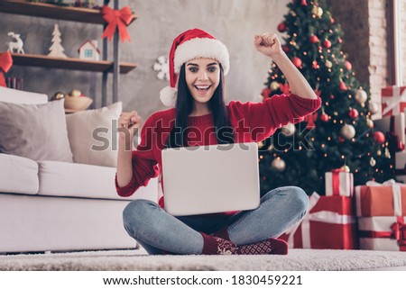 Photo of charming excited girl sit carpet hold laptop raise fists wear santa cap red pullover jeans socks in decorated living room indoors