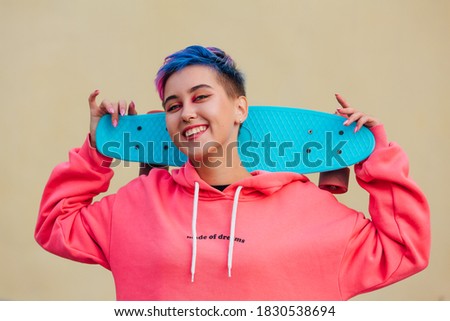 Stylish young woman with short colored hair holding blue plastic skateboard on her shoulders. Youth concept.