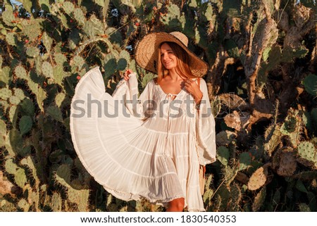 Young woman dressed in white dress and straw hat  standing among cactuses, smile and dance