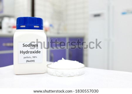 Selective focus of a bottle of pure sodium hydroxide or NaOH chemical compound beside a petri dish with white solid pellets. Chemistry research laboratory background with copy space. Royalty-Free Stock Photo #1830557030