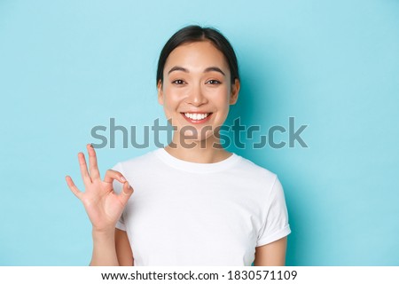 Close-up of pretty satisfied smiling asian girl in white t-shirt, showing recommendation gesture, okay sign and nod in approval, compliment choice, agree with you, standing light blue background