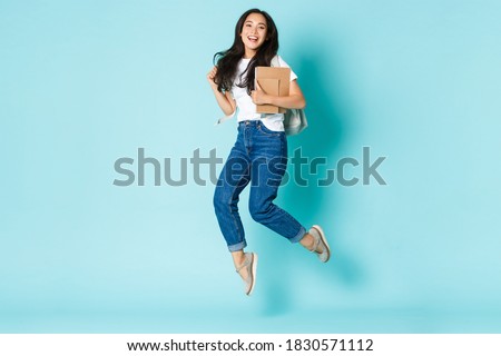 Fashion, back to school and lifestyle concept. Cheerful young asian girl, korean student looking upbeat, holding backpack and notebooks, jumping upbeat over light blue background