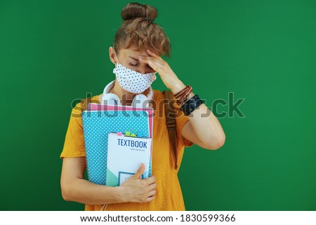 Life during covid-19 pandemic. stressed young student in yellow shirt with textbooks, notebooks, medical mask, white headphones and backpack isolated on green. Royalty-Free Stock Photo #1830599366