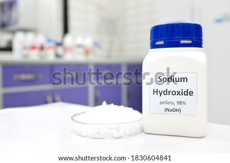 Selective focus of a bottle of pure sodium hydroxide or NaOH chemical compound beside a petri dish with white solid pellets. Chemistry research laboratory background with copy space. Royalty-Free Stock Photo #1830604841