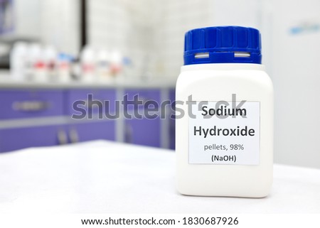 Selective focus of a bottle of pure sodium hydroxide or NaOH chemical compound beside a petri dish with white solid pellets. Chemistry research laboratory background with copy space. Royalty-Free Stock Photo #1830687926