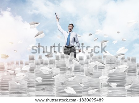 Businessman keeping hand with book up while standing among flying paper planes with cloudly skyscape on background. Mixed media.