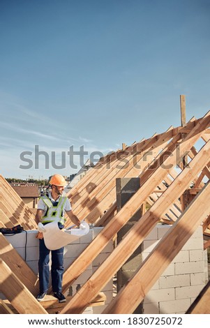 Full-length photo of a busy builder holding a huge blueprint and looking at the construction site