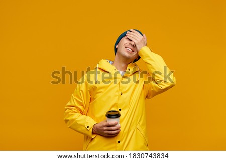 Portrait of positive cool man in warm autumn top outdoor clothes holding coffee and closing eyes with hands, male isolated over yellow color background. Bright casual wear, yellow cloak Royalty-Free Stock Photo #1830743834