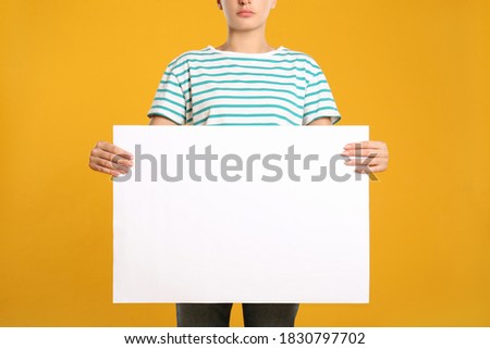 Woman holding white blank poster on yellow background, closeup. Mockup for design