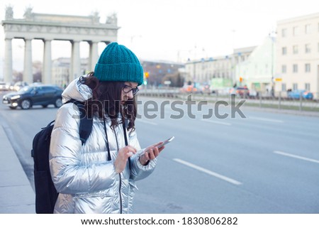woman in a down jacket and hat stands at a public transport stop and uses a smartphone. Communication in messengers and social networks, mobile Internet