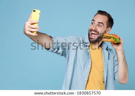 Funny happy young bearded man 20s in casual clothes posing holding in hands american classic fast food burger doing selfie shot on mobile phone isolated on pastel blue color background studio portrait