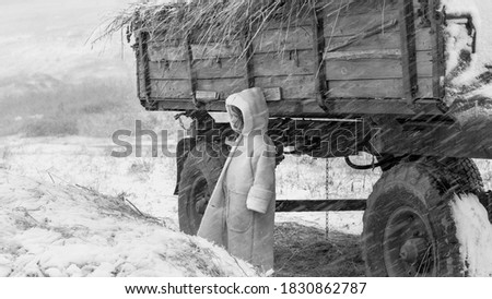 А little girl in the winter in a warm coat hiding from the snow blizzard behind a big car. Image with selective focus. Black and white image. Focus on the girl and car.