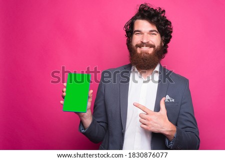 Happy bearded man wearing suit and pointing at green screen on tablet near pink background