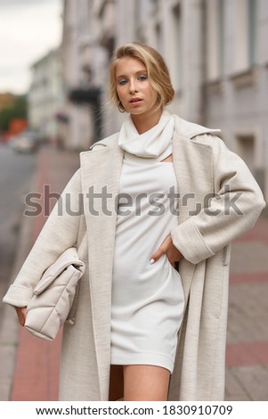 Stylish elegant girl in white coat and dress. Pretty young woman with makeup and hairstyle at city street. Autumn fashion. Monochrome look