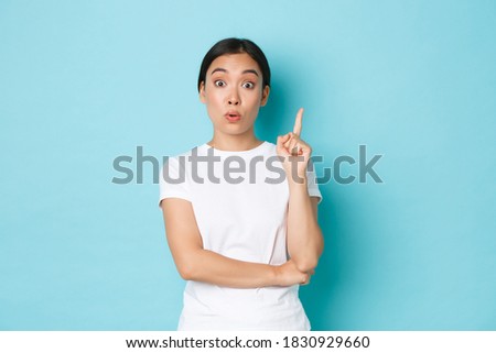 Lifestyle, beauty and shopping concept. Excited asian girl in white t-shirt, raising index finger, have suggestion, think-up plan or solution, saying plan, standing blue background